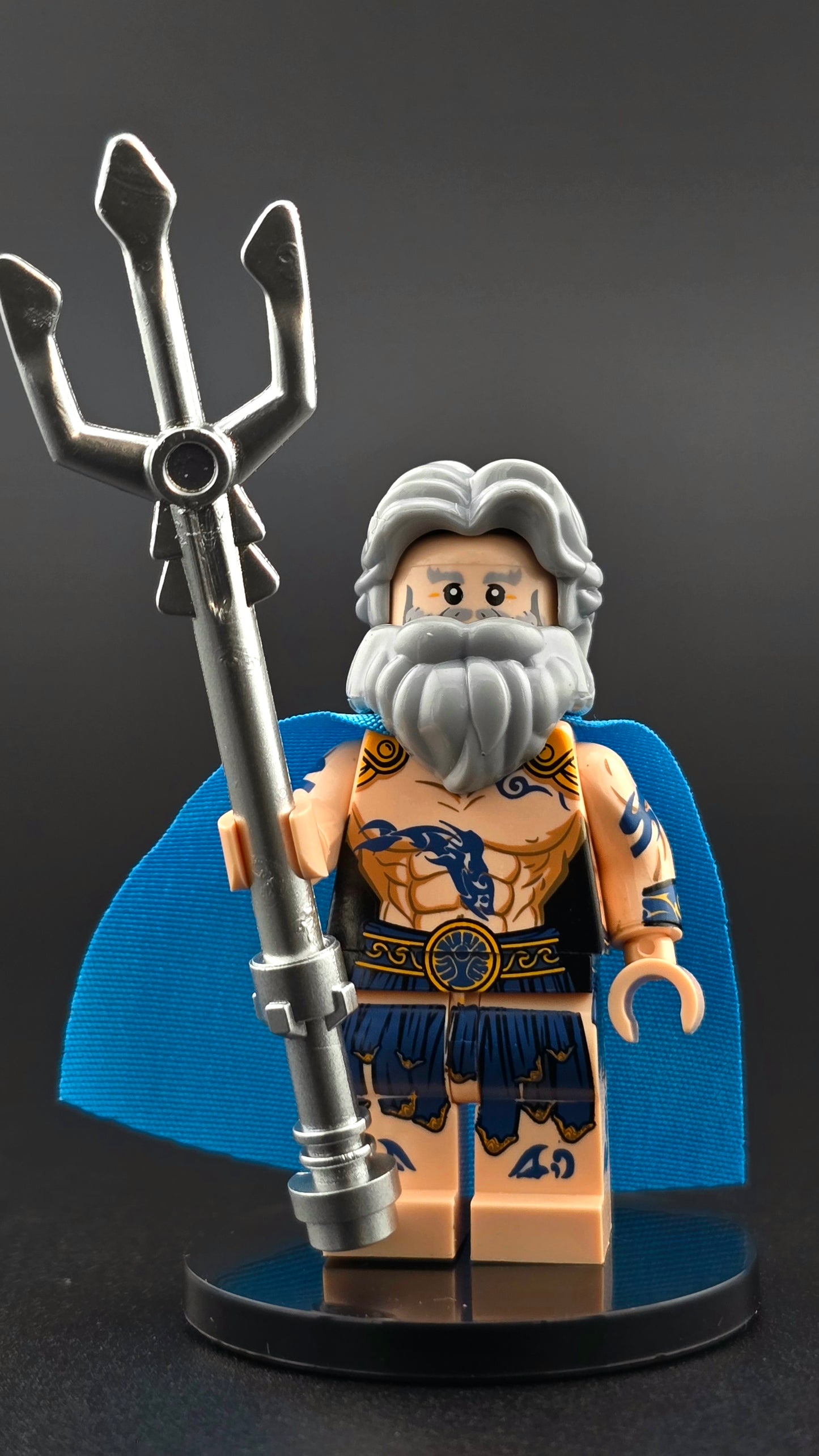 Custom Poseidon inspired minifigure with gray hair, beard, blue tattoos, and a flowing blue cape, holding a silver trident. Detailed print and compatible with leading brick-style building systems.