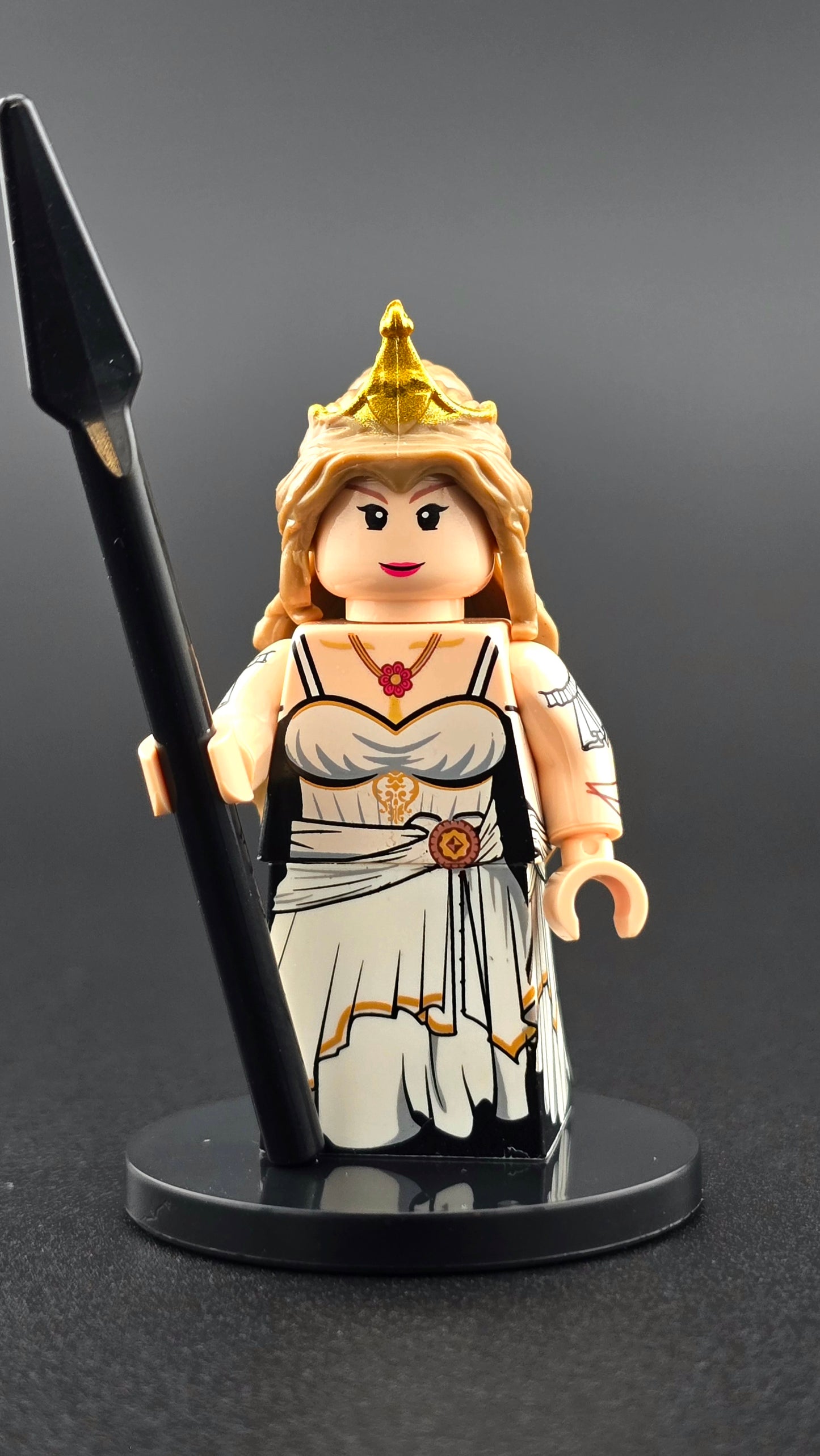 Custom mythology-inspired Hera minifigure in a white and gold regal toga with detailed front and back printing, shown with spear and interchangeable helmet and tiara hairpiece. Premium custom brick figure compatible with major brick-style building systems.