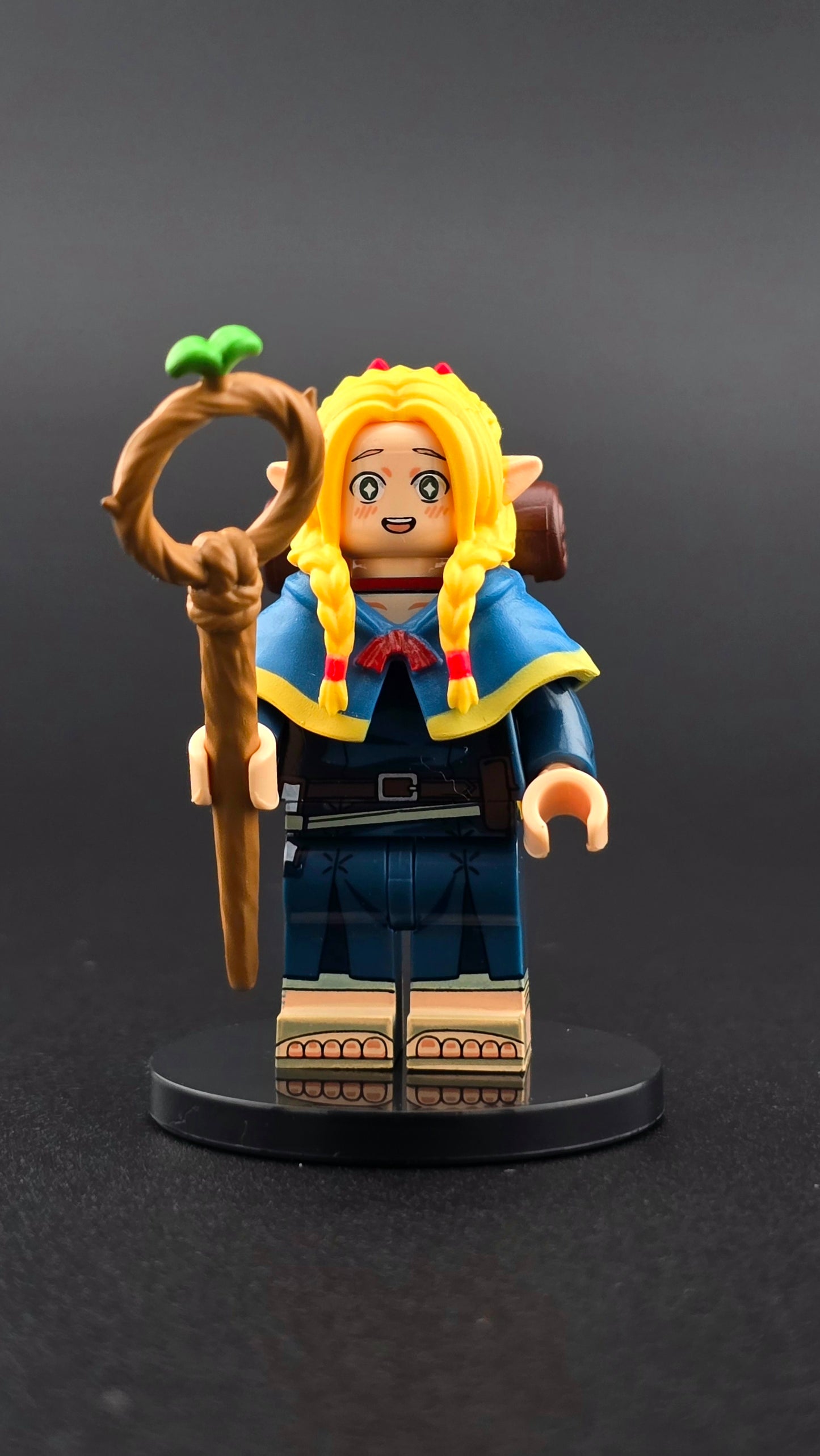 Custom anime inspired Marcille minifigure with blue and yellow mage outfit, blonde hair, dual sided head, staff, food bowl, chopsticks, and backpack. Designed with detailed fantasy styling compatible with leading brick style building sets.