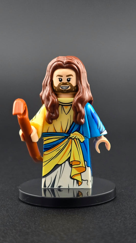 Custom religion-inspired Jesus minifigure wearing a white toga with blue and yellow accents, smiling with brown hair and holding a detailed staff. Designed for adult collectors and compatible with major brick-style systems.