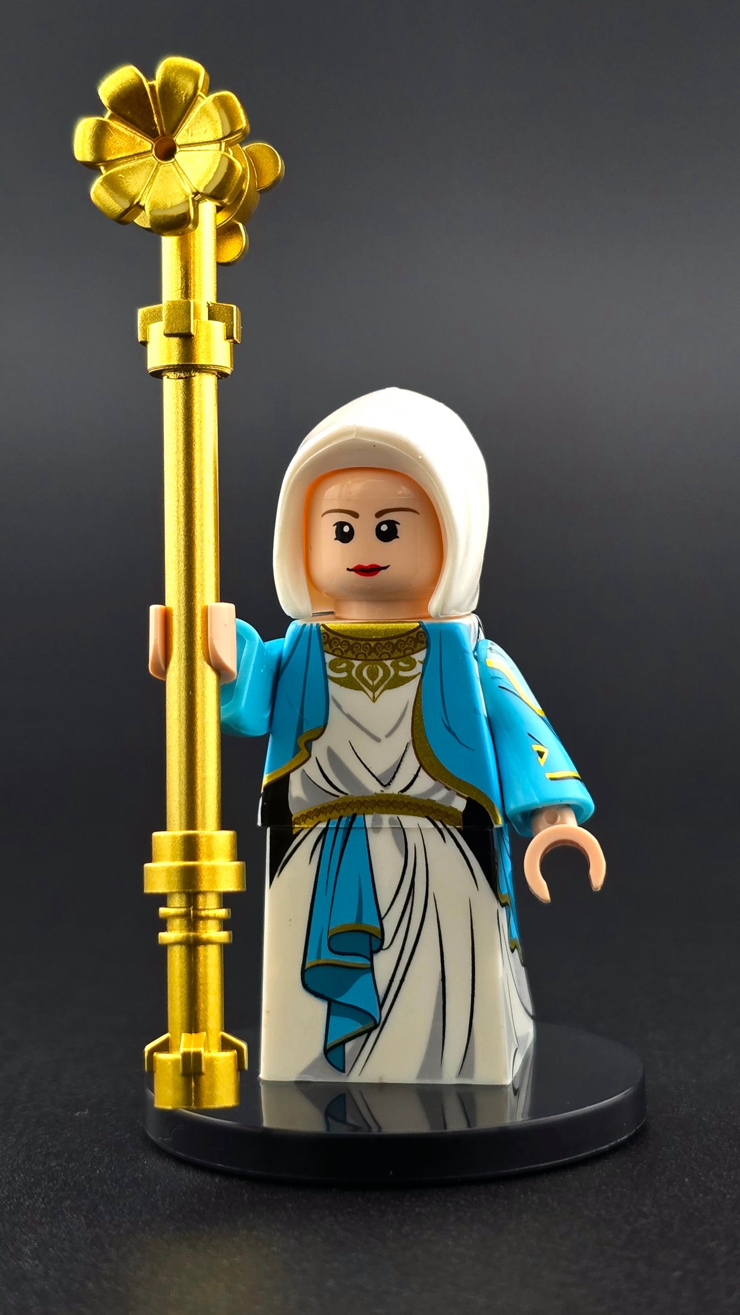 Custom Virgin Mary inspired minifigure in white and blue robes with gold details, featuring a compassionate expression and holding a golden staff. High quality print and compatible with leading brick-style building sets.