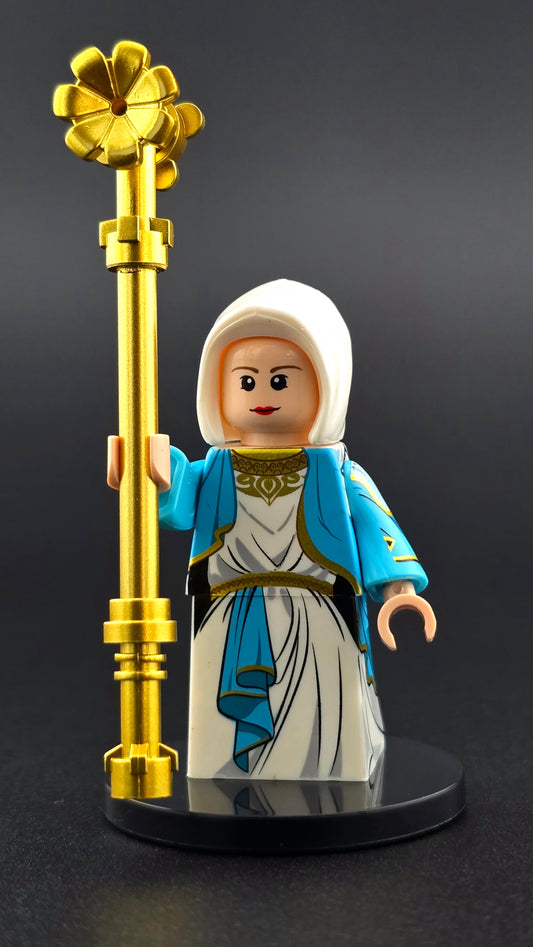 Custom Virgin Mary inspired minifigure in white and blue robes with gold details, featuring a compassionate expression and holding a golden staff. High quality print and compatible with leading brick-style building sets.
