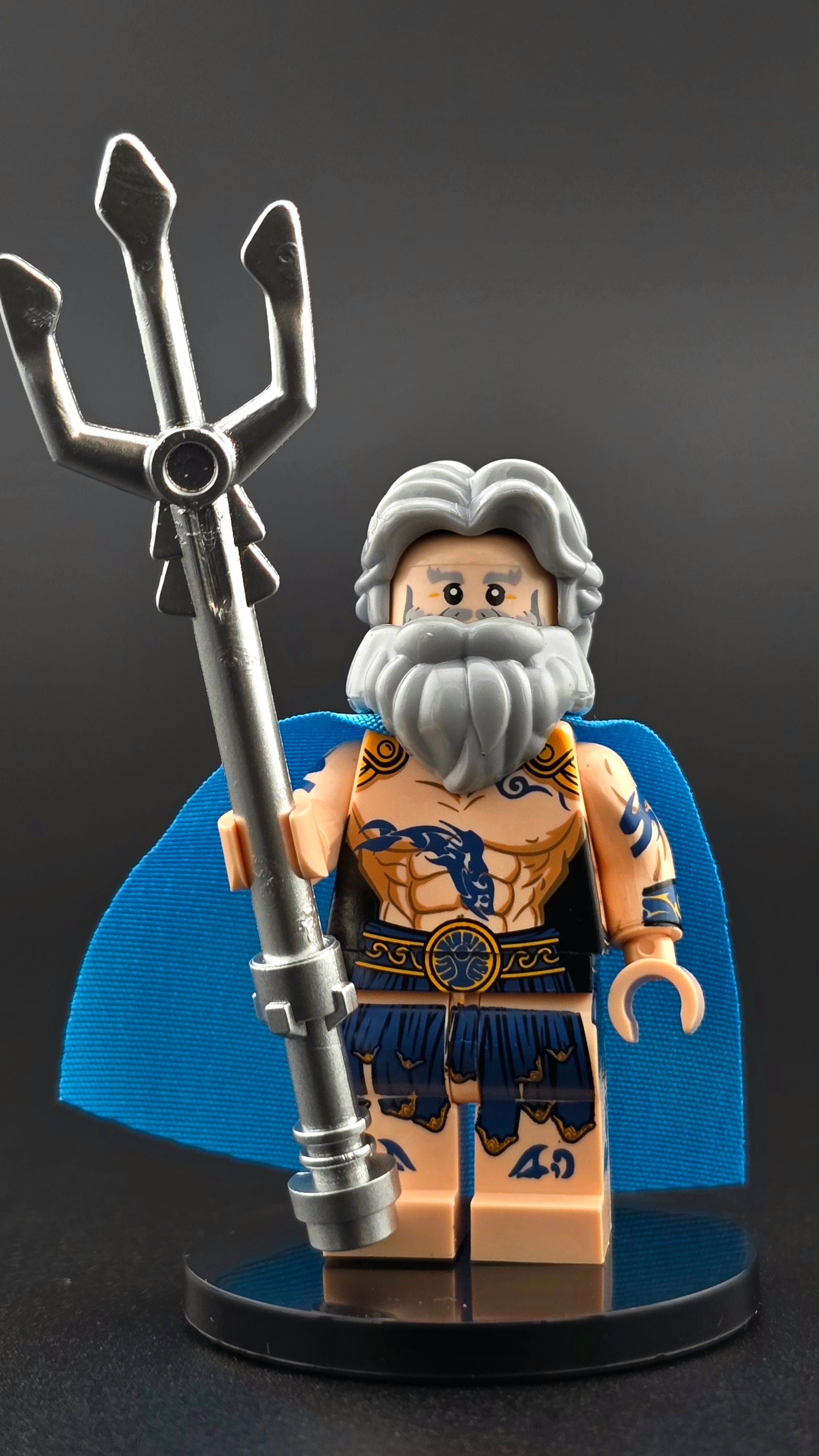 Custom Poseidon inspired minifigure with gray hair, beard, blue tattoos, and a flowing blue cape, holding a silver trident. Detailed print and compatible with leading brick-style building systems.