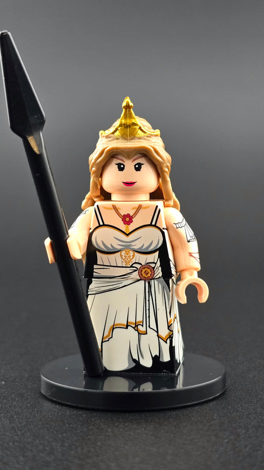 Custom mythology-inspired Hera minifigure in a white and gold regal toga with detailed front and back printing, shown with spear and interchangeable helmet and tiara hairpiece. Premium custom brick figure compatible with major brick-style building systems.