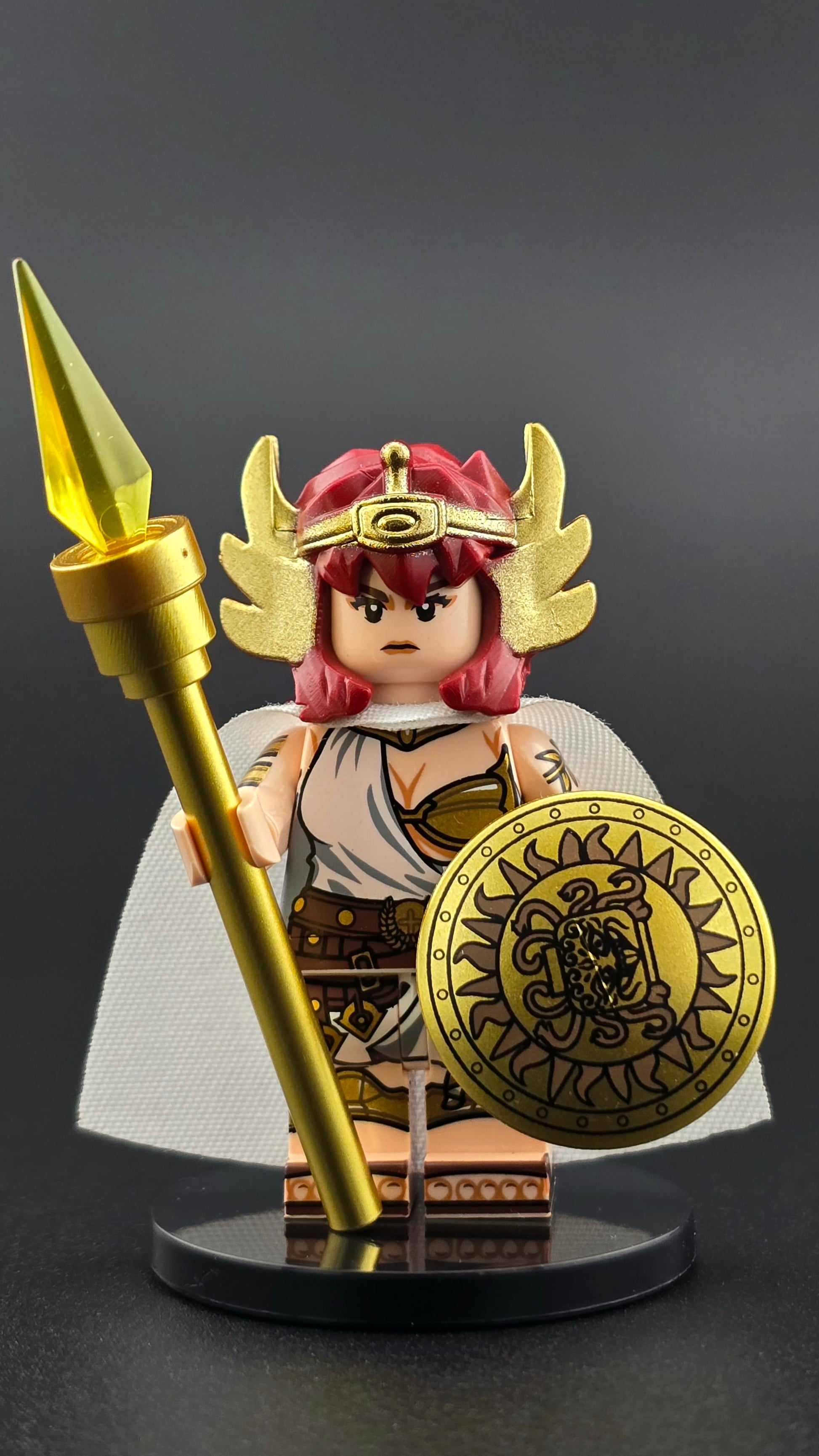 Custom mythology-inspired Athena minifigure with red hair, winged headgear, white and gold toga armor, golden spear, and ornate shield. Detailed front printing and premium accessories compatible with major brick-style building systems.