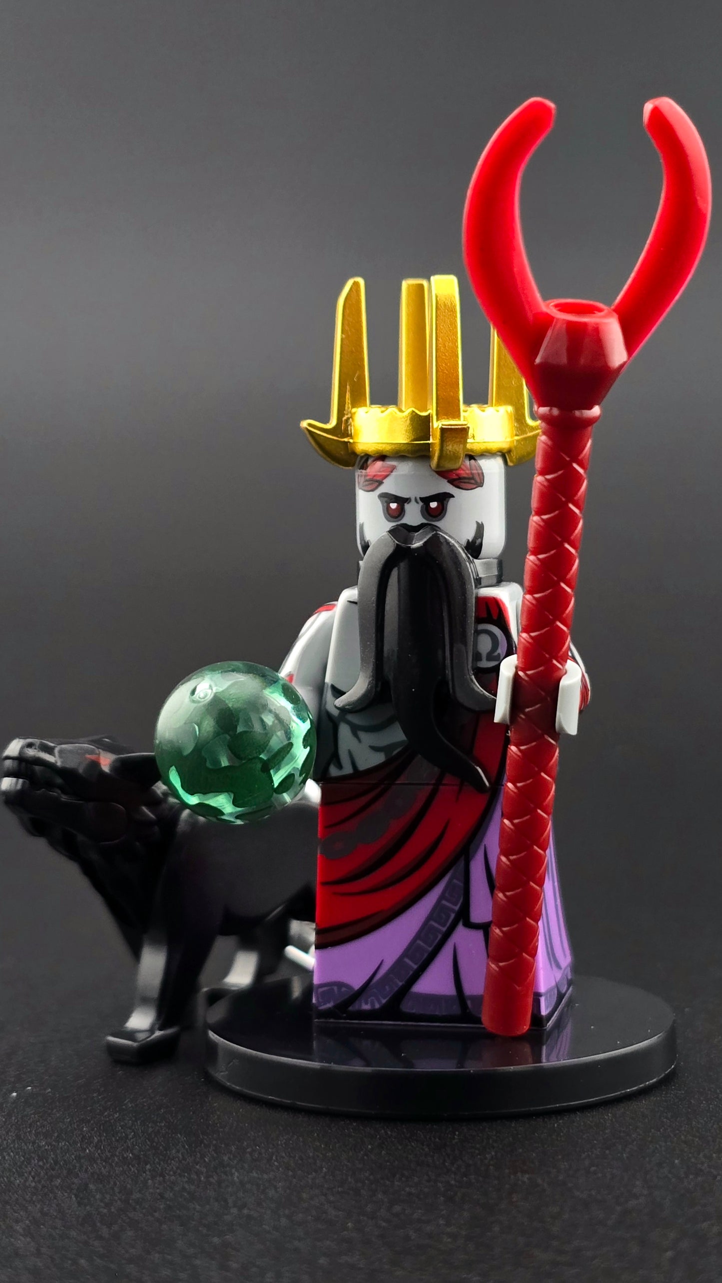 Custom mythology-inspired Hades minifigure wearing a red and purple toga with a golden crown, black beard, and red eyes, holding a red staff and green orb. Includes a black hound companion and features bold red, purple, black, and gold details compatible with leading brick-style sets.