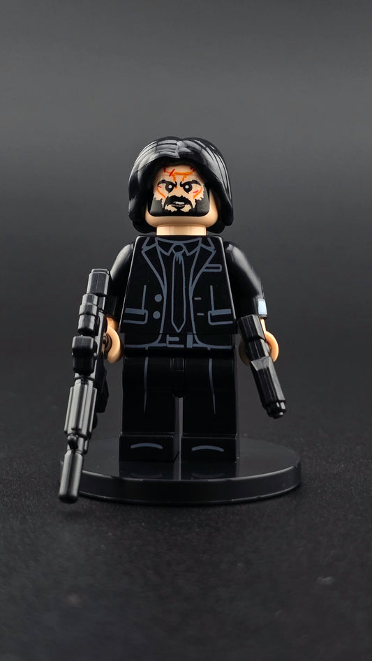 Custom action movie inspired hitman minifigure in an all black suit with bruised face, holding an assault rifle and pistol. Premium painted details and compatible with leading brick style building sets.