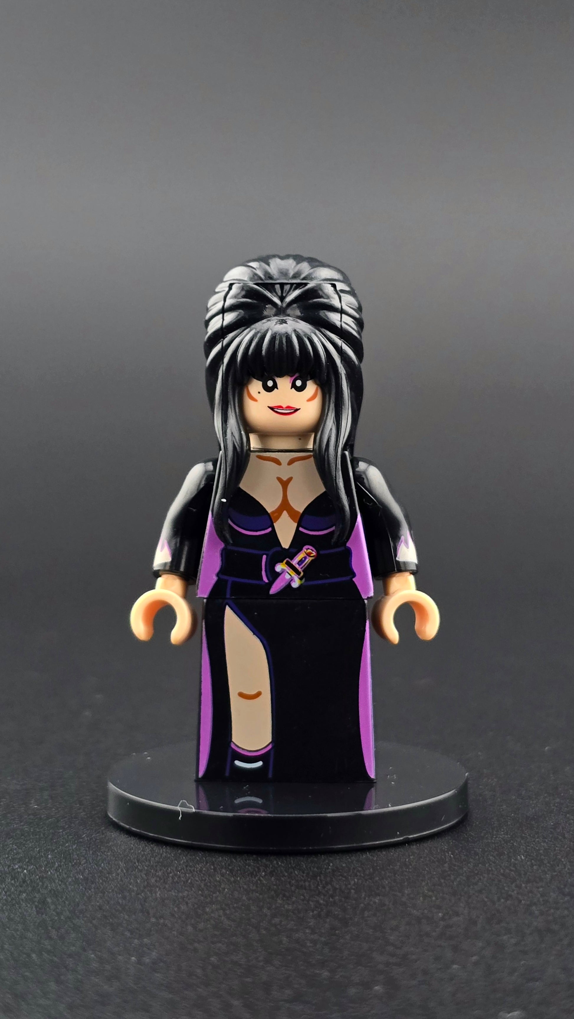 Custom movie inspired Elvira style minifigure with big black hair, smirking expression, and black and purple dress. Detailed gothic themed design compatible with leading brick style building sets.