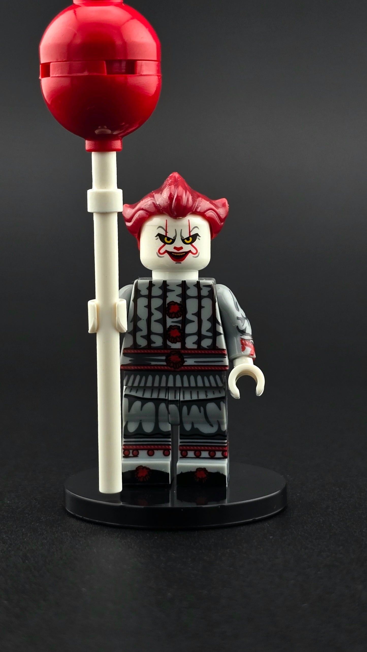 Custom horror inspired Pennywise style minifigure in gray and red clown outfit with sinister grin and red balloon accessory. Detailed creepy themed design compatible with leading brick style building sets.
