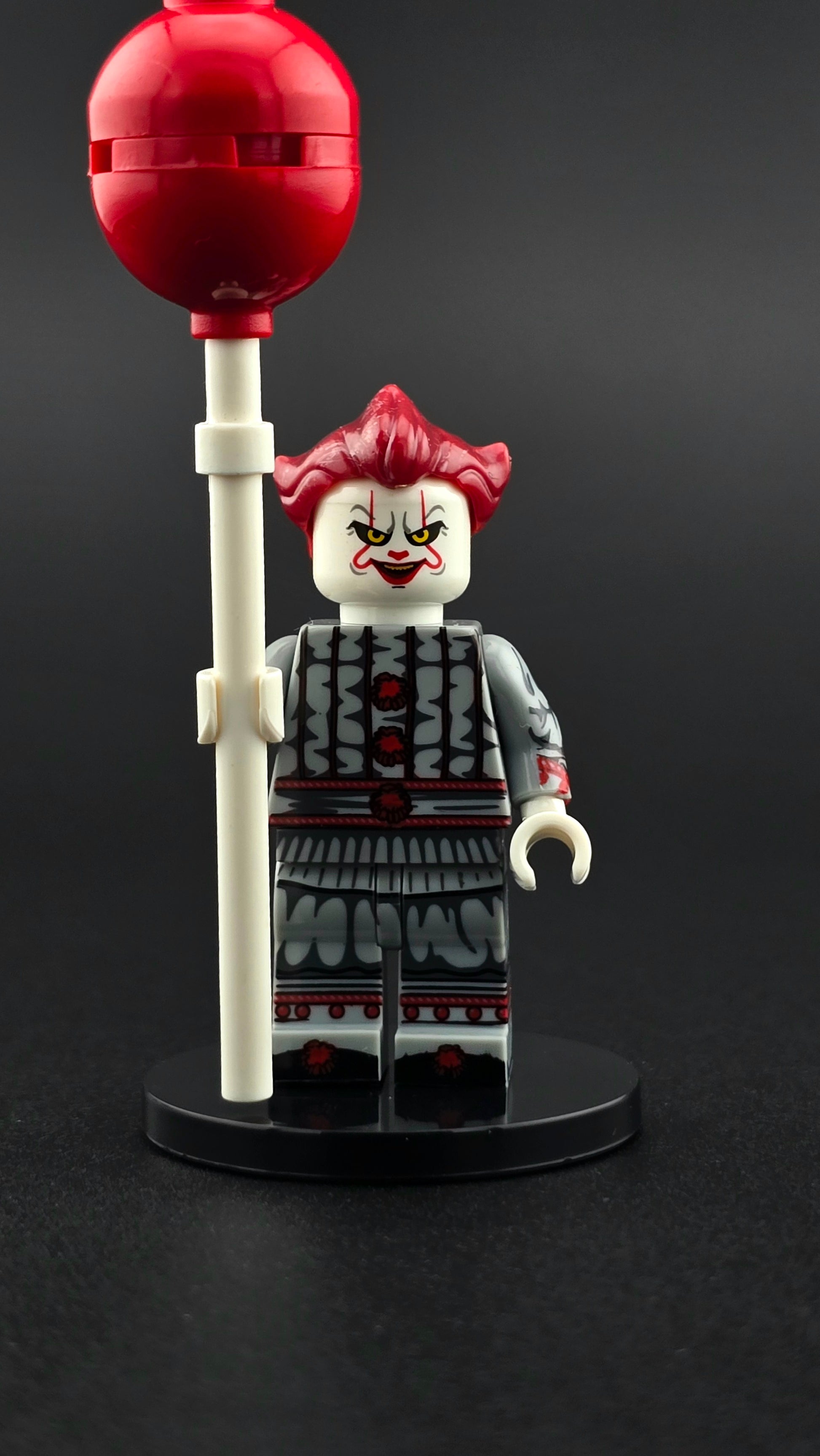 Custom horror inspired Pennywise style minifigure in gray and red clown outfit with sinister grin and red balloon accessory. Detailed creepy themed design compatible with leading brick style building sets.