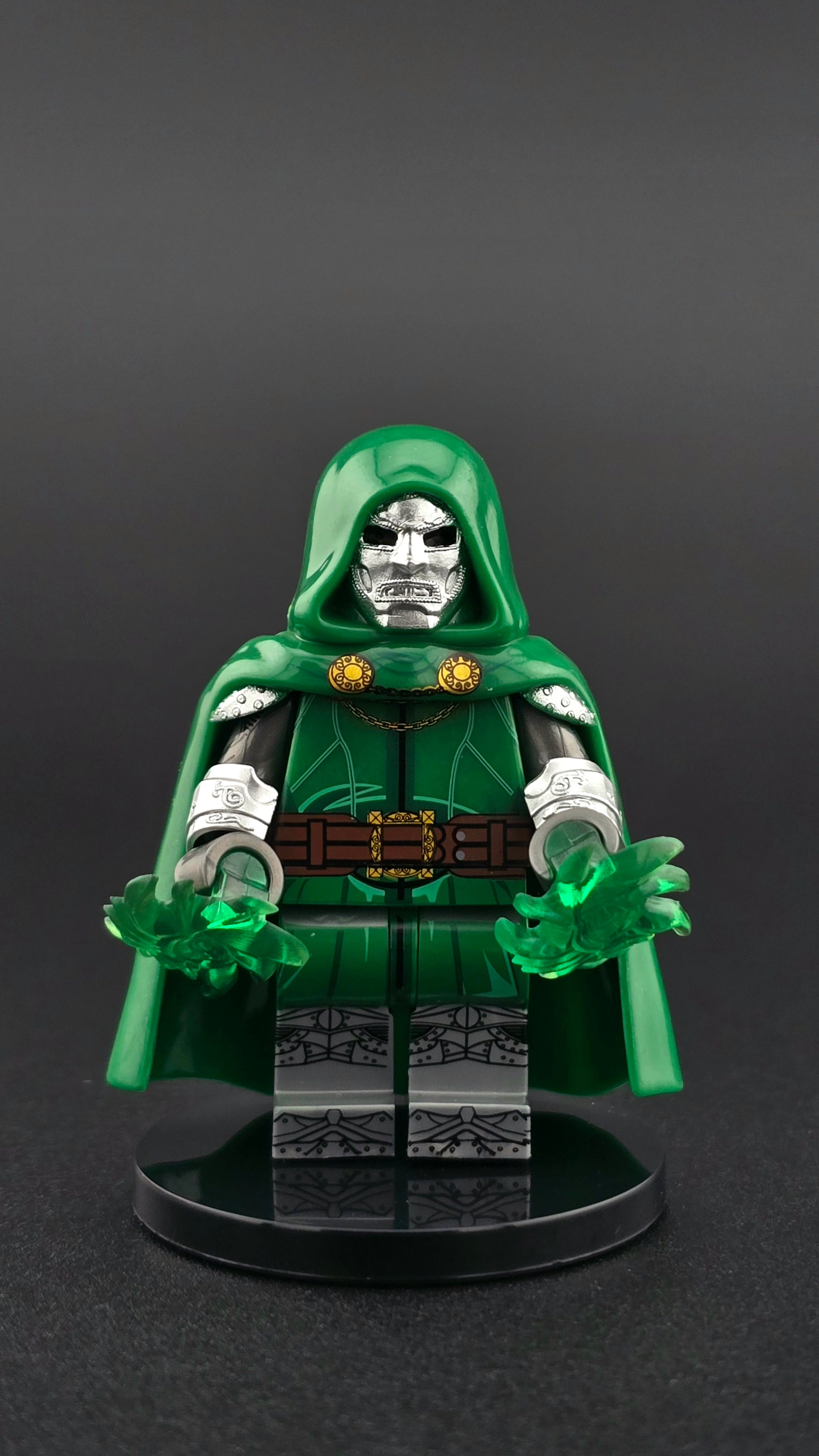 Custom villain-inspired Dr. Doom minifigure with silver metal mask, green hood and cape, dual-sided head, and green power energy accessory, compatible with leading brick-style building systems.
