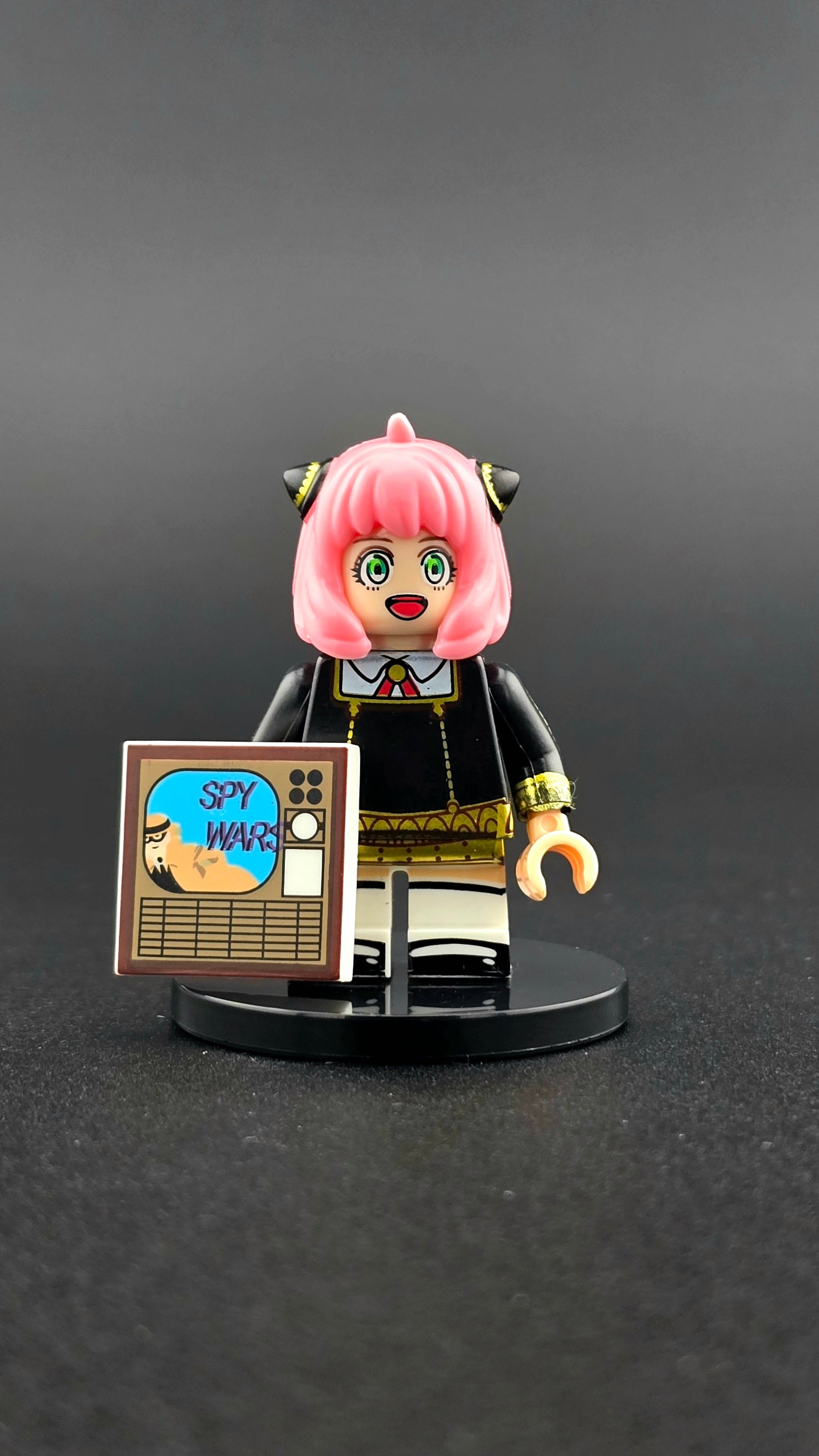 Custom anime-inspired Anya Forger minifigure with pink hair, school outfit, and Spy Wars TV cover accessory, based on Spy X Family and compatible with leading brick-style building systems.